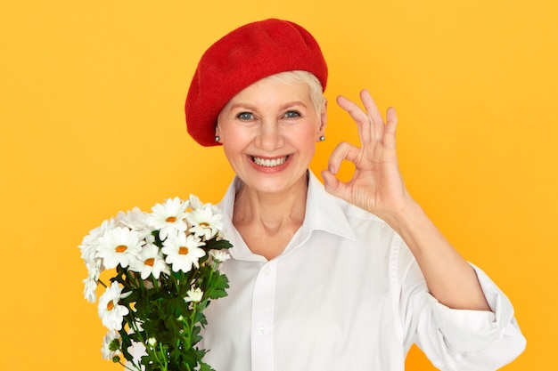 Retrato de uma florista atraente, alegre, de meia-idade, com chapéu vermelho, tendo uma expressão facial confiante, fazendo um gesto certo, segurando um monte de margaridas, organizando flores para um evento especial