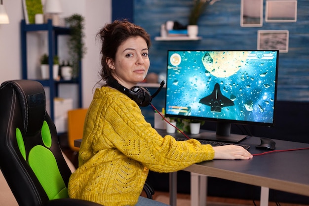 Retrato de uma gamer mulher sentada na mesa jogando videogame de atirador espacial durante o campeonato online usando RGB computador poderoso na sala de estar em casa. Torneio Esport do estúdio de jogo