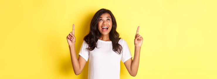 Foto grátis retrato de uma garota africana feliz espantada sorrindo e apontando dedos para cima em pé sobre b amarelo