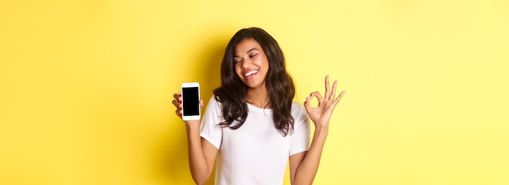 Foto grátis retrato de uma garota afro-americana bonita sorrindo satisfeita mostrando um sinal de ok e a tela do telefone móvel reco