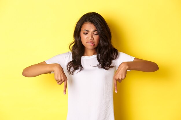 Retrato de uma garota afro-americana duvidosa e cética em camiseta branca, franzindo a testa e apontando os dedos para algo estranho ou desagradável, em pé sobre um fundo amarelo.