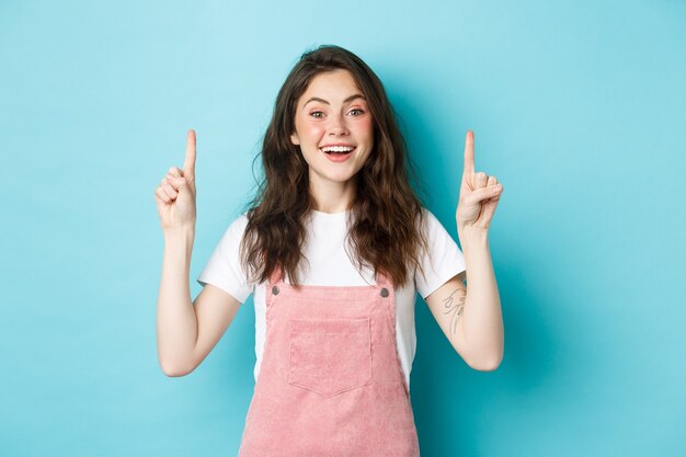 Retrato de uma garota alegre e glamour, sorrindo feliz, parecendo animada e apontando o dedo para cima, mostrando um anúncio, vestindo roupas de verão, fundo azul