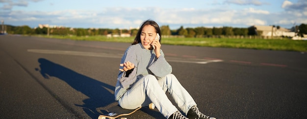 Retrato de uma garota asiática sentada em seu longboard patinando em skateboard e falando em celular