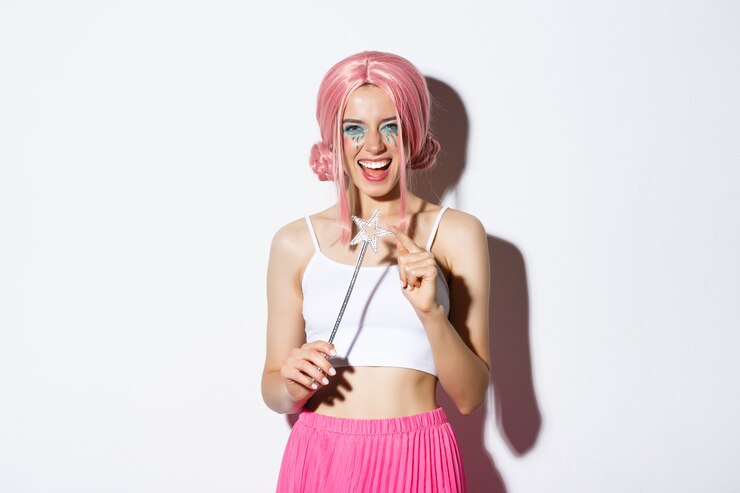 Foto grátis retrato de uma garota atraente com peruca rosa e maquiagem brilhante, vestida de fada para a festa de halloween, segurando uma varinha mágica e sorrindo