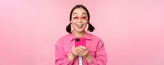 Retrato de uma garota coreana em óculos de sol elegantes, segurando o celular usando o aplicativo de smartphone sobre fundo rosa