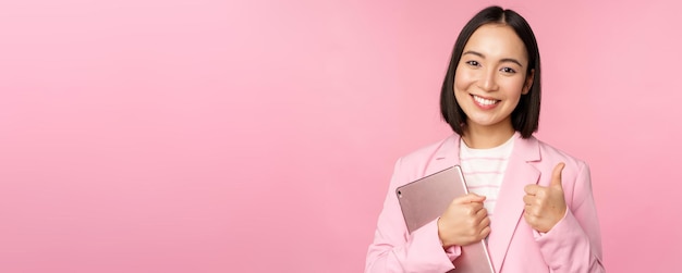Retrato de uma garota corporativa no escritório em um terno de negócios segurando o tablet digital mostrando os polegares