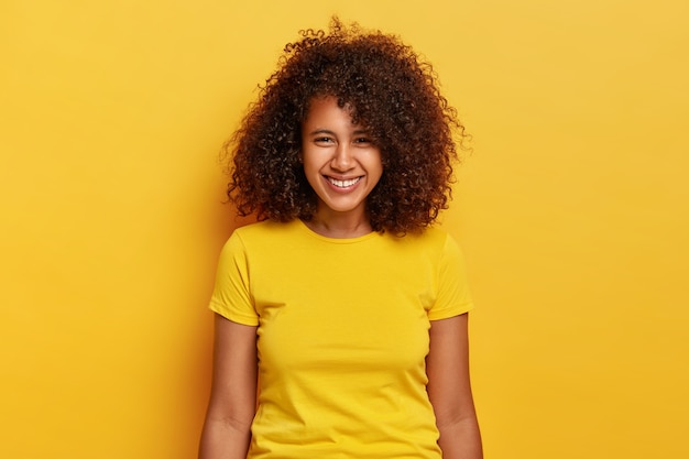 Retrato de uma garota feliz e hippie ri positivamente, ri e sorri despreocupado, usa roupas casuais amarelas brilhantes, tem pouco espaço entre os dentes, sente-se encantado, posa dentro de casa, tem beleza natural