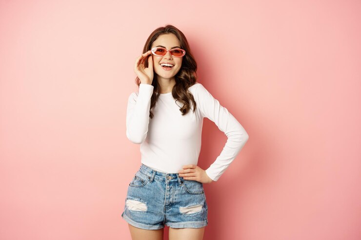 Foto grátis retrato de uma garota glam elegante com óculos de sol rindo e sorrindo de pé sobre um fundo rosa de férias