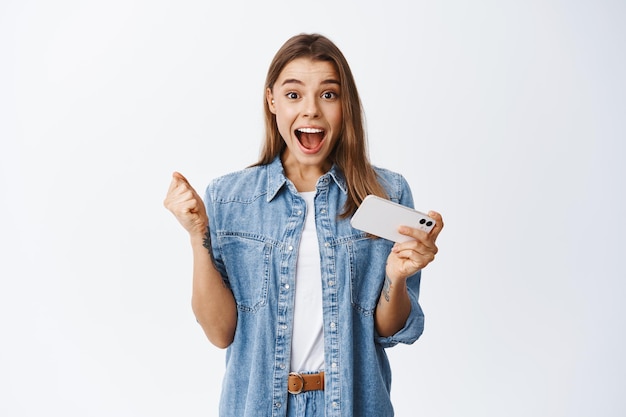 Retrato de uma garota loira animada ganhando dinheiro ou prêmio online, segurando o telefone celular e gritando de alegria, comemorando a vitória, vencendo no videogame no smartphone, branco