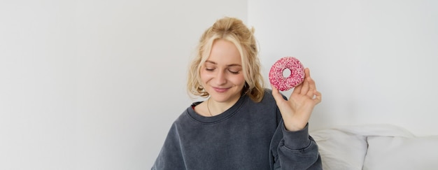 Foto grátis retrato de uma garota loira bonita segurando um donut rosa com salpicaduras em cima mostrando sua comida favorita