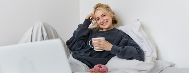 Retrato de uma garota loira feliz, modelo feminina, deitada na cama, com roupas confortáveis, comendo e vendo um filme.