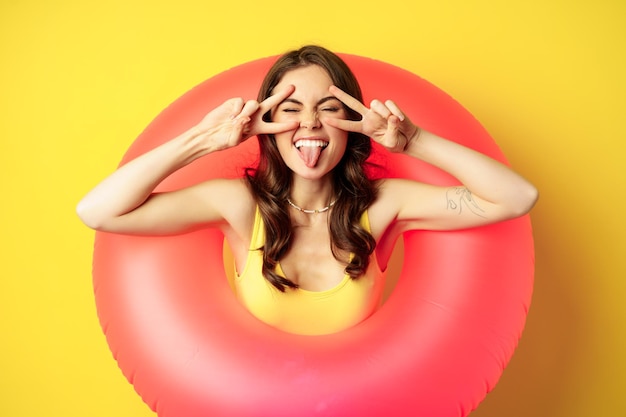 Foto grátis retrato de uma garota moderna atraente dentro do anel de natação na moda praia rosa, mostrando paz v-sign e sorrindo feliz, de pé sobre fundo amarelo