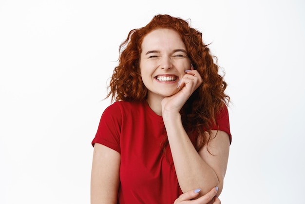 Retrato de uma garota super feliz com cabelo ruivo, regozijando-se com os olhos fechados e sorrindo alegremente recebe ótimas notícias em pé otimista contra um fundo branco em camiseta casual