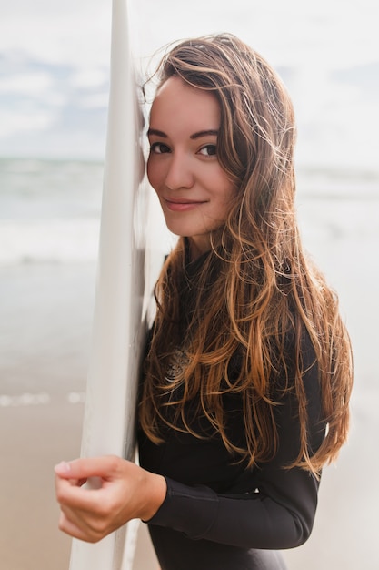 Retrato de uma jovem adorável e fofa com longos cabelos castanhos-claros vestida com fantasia de surf e segurando uma prancha de surf no fundo do oceano e areia branca, estilo de vida esportivo ativo