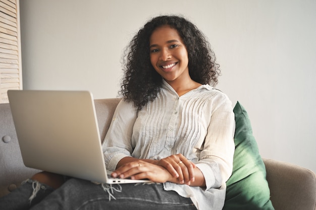 Foto grátis retrato de uma jovem africana alegre em jeans e camiseta, sorrindo amplamente enquanto navega na internet em um computador portátil genérico, desfrutando de uma conexão sem fio de alta velocidade na sala de estar
