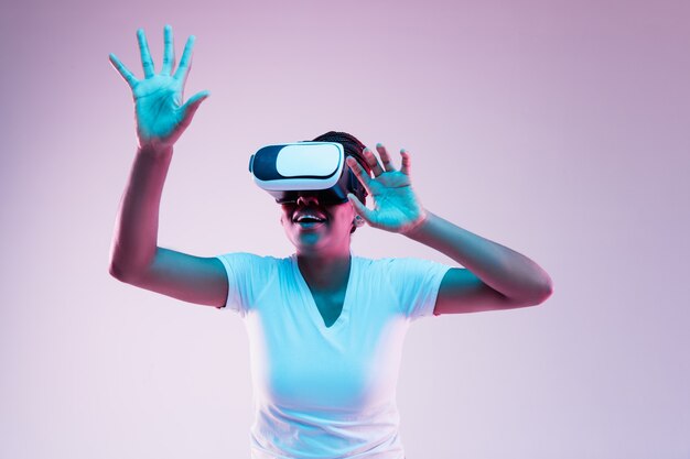 Retrato de uma jovem afro-americana brincando com óculos VR em luz de néon em fundo gradiente