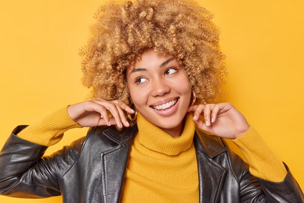 Retrato de uma jovem alegre e despreocupada desvia o olhar com expressão feliz, gosta de seus novos brincos, sente sorrisos alegres, usa um macacão casual e jaqueta de couro isolada sobre fundo amarelo