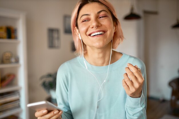 Retrato de uma jovem alegre e feliz com cabelo rosa e piercing no nariz, posando na elegante sala de estar usando fones de ouvido