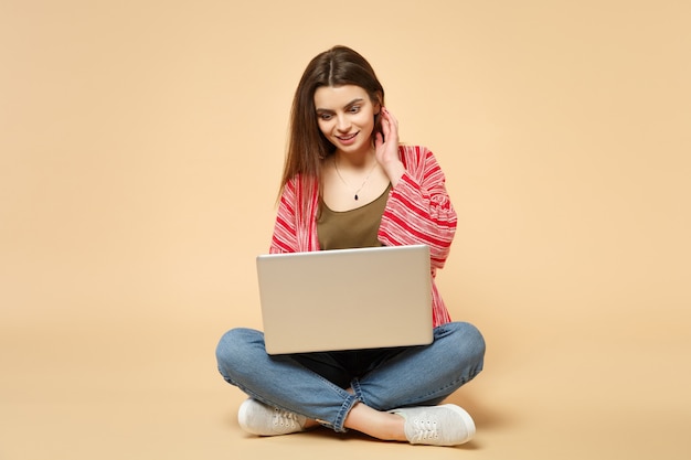 Retrato de uma jovem animada em roupas casuais, sentado, usando o computador laptop pc isolado no fundo da parede bege pastel no estúdio. conceito de estilo de vida de emoções sinceras de pessoas. simule o espaço da cópia.