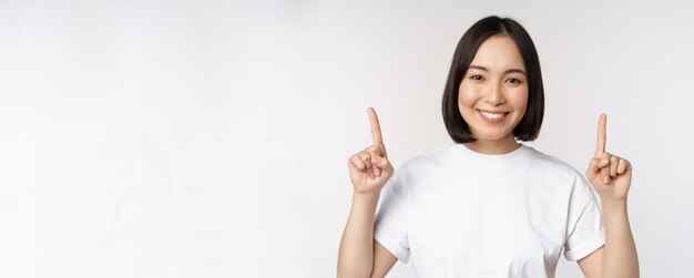 Retrato de uma jovem asiática entusiasmada sorrindo apontando os dedos para cima, mostrando o anúncio