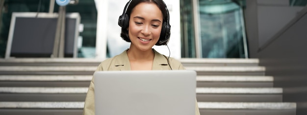 Foto grátis retrato de uma jovem asiática sentada com laptop e fones de ouvido assistindo vídeo faz curso on-line