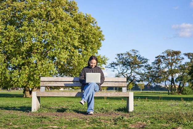 Foto grátis retrato de uma jovem asiática trabalhando em um laptop sentado no banco do parque em um dia ensolarado usando seu comp