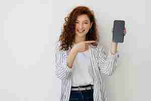 Foto grátis retrato de uma jovem atraente com cabelos ruivos longos e encaracolados isolados sobre fundo branco mostrando o celular em branco