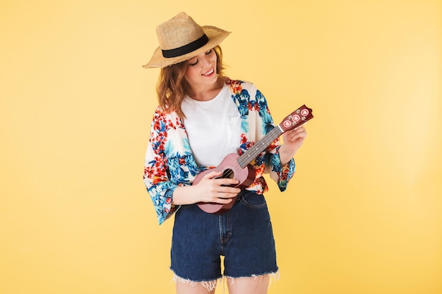 Foto grátis retrato de uma jovem bonita de chapéu de palha e camisa colorida em pé com um pequeno violão e alegremente tocando sobre fundo rosa