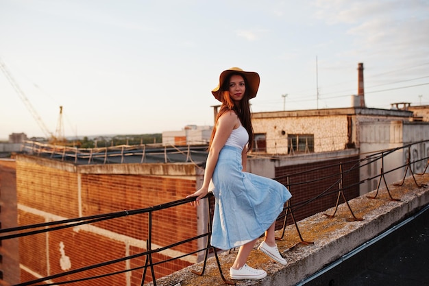 Retrato de uma jovem bonita em camiseta branca e saia azul posando no telhado com seu chapéu laranja ao pôr do sol