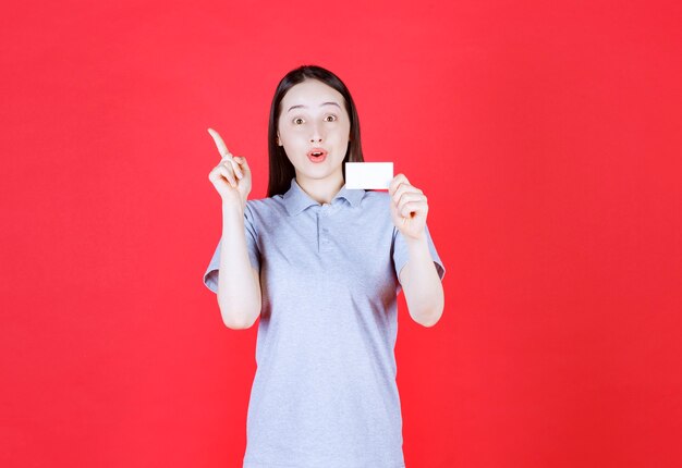 Retrato de uma jovem bonita segurando seu cartão de visita e apontando o dedo para cima