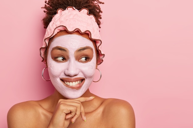 Retrato de uma jovem bonita tocando o queixo, com um sorriso gentil e cheio de dentes, aplicando máscara de argila para rejuvenescimento, ombros nus encostados na parede rosa