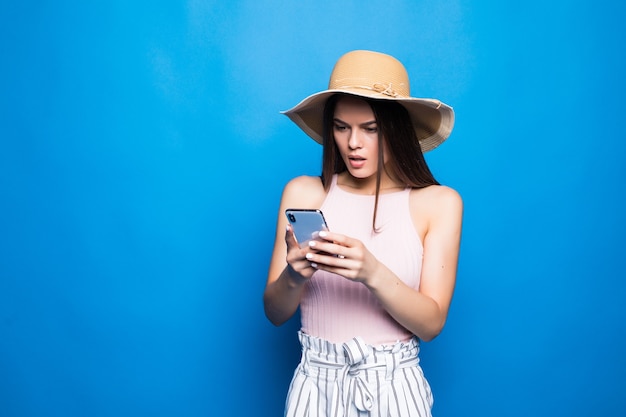 Retrato de uma jovem chocada com chapéu de verão, olhando para o celular isolado sobre a parede azul.