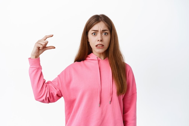 Retrato de uma jovem confusa mostrando um gesto minúsculo olhando nervoso para a câmera exibir um pequeno objeto com rosto ansioso sobre fundo branco