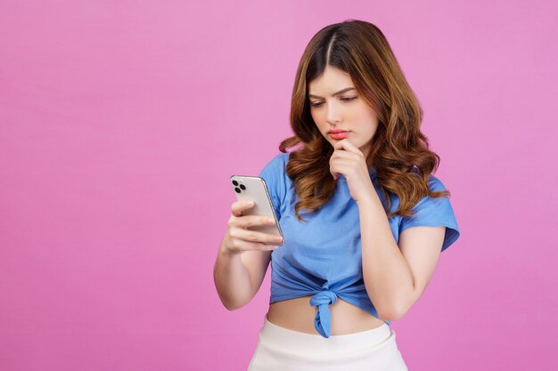 Retrato de uma jovem confusa vestindo camiseta casual usando smartphone isolado sobre fundo rosa