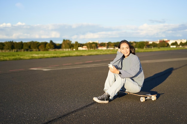 Retrato de uma jovem coreana sentada em seu skate na estrada olhando para smartphone conversando em mo