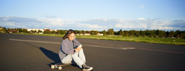 Foto grátis retrato de uma jovem coreana sentada em seu skate na estrada olhando para um smartphone conversando