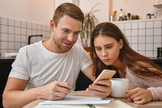 Retrato de uma jovem curiosa focada, sentada à mesa da cozinha e lendo notícias positivas no telefone nas mãos do marido. Homem atraente preenchendo documentos financeiros, usando aplicativo de banco on-line via celular