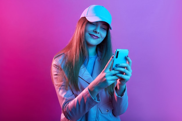 Retrato de uma jovem e bela modelo elegante usando couro padeiro e boné de beisebol, segurando o telefone inteligente nas mãos