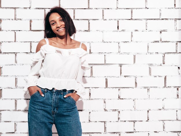 Retrato de uma jovem e linda mulher negra Modelo sorridente vestido com roupas de jeans de verão Mulher despreocupada sexy posando perto da parede de tijolos brancos no estúdio Bronzeado e alegre