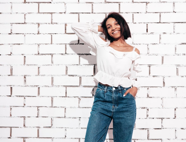 Foto grátis retrato de uma jovem e linda mulher negra modelo sorridente vestido com roupas de jeans de verão mulher despreocupada sexy posando perto da parede de tijolos brancos no estúdio bronzeado e alegre
