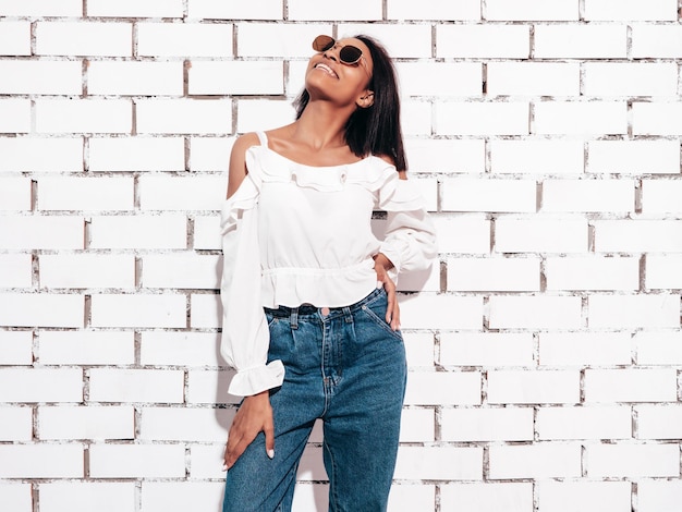 Foto grátis retrato de uma jovem e linda mulher negra modelo sorridente vestido com roupas de jeans de verão mulher despreocupada sexy posando perto da parede de tijolos brancos no estúdio bronzeado e alegre