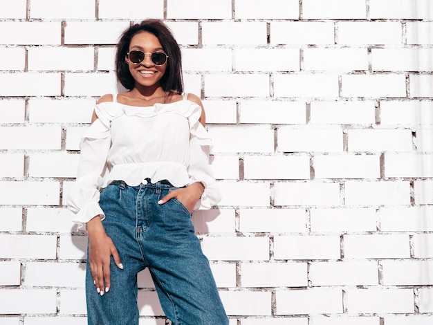 Retrato de uma jovem e linda mulher negra Modelo sorridente vestido com roupas de jeans de verão Mulher despreocupada sexy posando perto da parede de tijolos brancos no estúdio Bronzeado e alegre