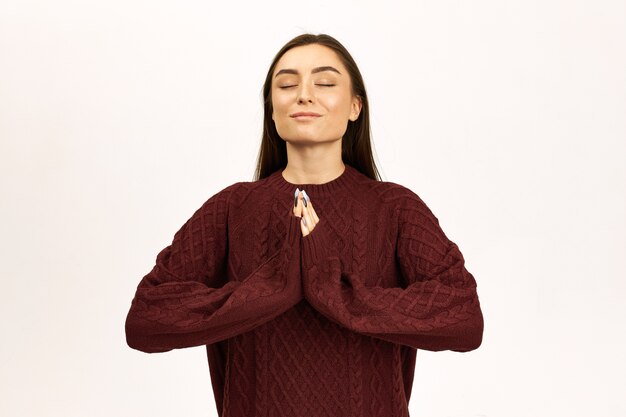 Retrato de uma jovem encantadora com cabelo comprido escuro posando em jumper, gesticulando, pressionando as palmas das mãos juntas, mantendo os olhos fechados, tendo uma expressão facial calma e pacífica, orando, sendo grato