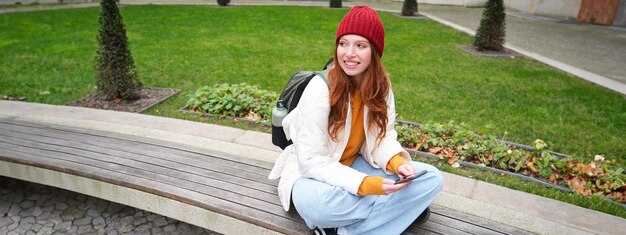 Retrato de uma jovem estilosa sentada no banco do parque e usando telefone celular lê notícias on-line