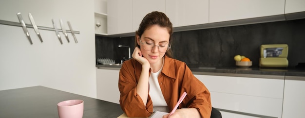 Foto grátis retrato de uma jovem estudante fazendo sua lição de casa estudando em casa sentada na cozinha fazendo anotações