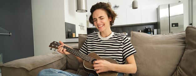 Retrato de uma jovem estudante moderna tocando ukulele em casa sentada com uma pequena guitarra cantando e