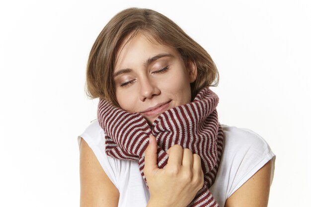 Retrato de uma jovem europeia atraente e satisfeita com maquiagem natural, fechando os olhos e sorrindo alegremente, desfrutando de um lenço de lã macio, se aquecendo em um dia frio de inverno