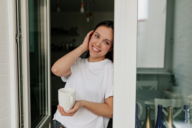 Retrato de uma jovem europeia com cabelo escuro e pele saudável, sorrindo com café da manhã na cozinha moderna e clara.