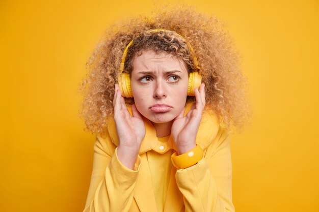 Retrato de uma jovem europeia de cabelos cacheados descontente mantém as mãos nos fones de ouvido e franze os lábios da expressão do rosto frustrado