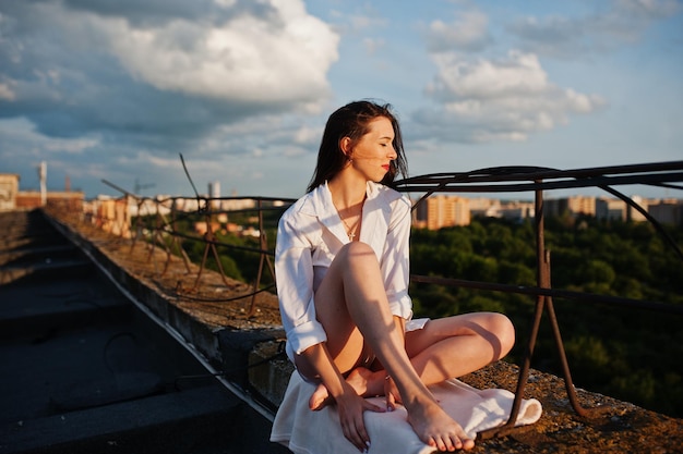 Foto grátis retrato de uma jovem fabulosa na camisa masculina branca, sentado na borda do telhado do edifício com uma vista fantástica de uma cidade ao fundo